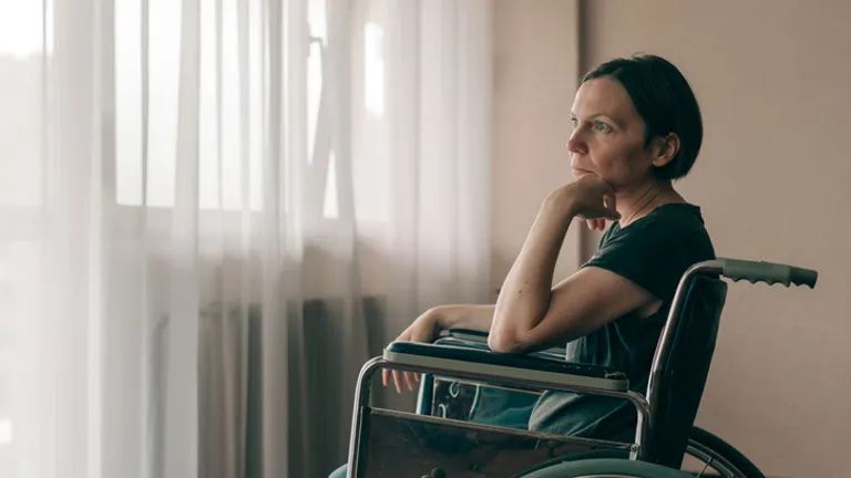 Person in a wheelchair by a sunlit window, resting their chin on their hand.