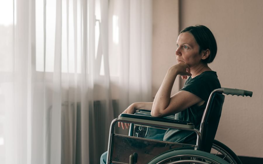 Person in a wheelchair by a sunlit window, resting their chin on their hand.