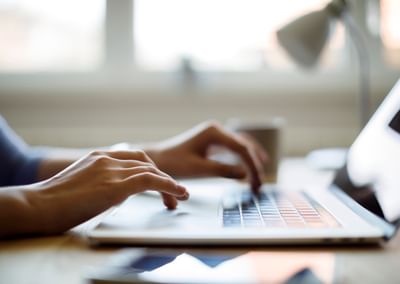 Person typing on a laptop at a desk, with a mug and a lamp in a blurred background.