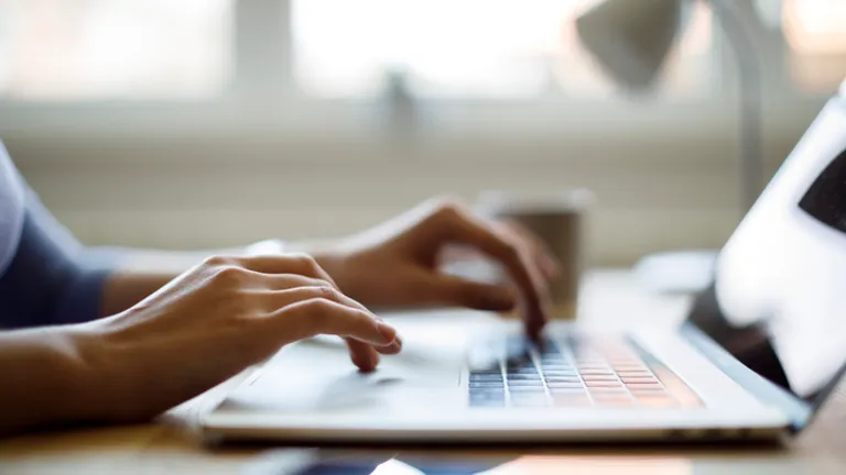 Person typing on a laptop at a desk, with a mug and a lamp in a blurred background.