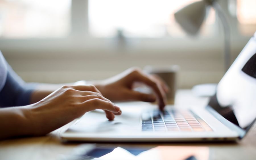 Person typing on a laptop at a desk, with a mug and a lamp in a blurred background.