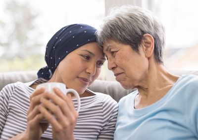 Two people sit close on a couch, heads touching; one wears a navy headscarf, the other in light blue.