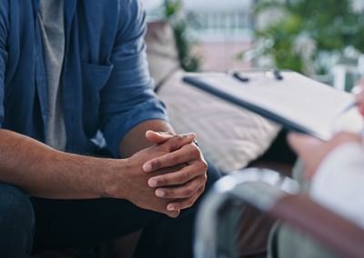 Person in blue shirt sits with clasped hands during a therapy session as another person takes notes.