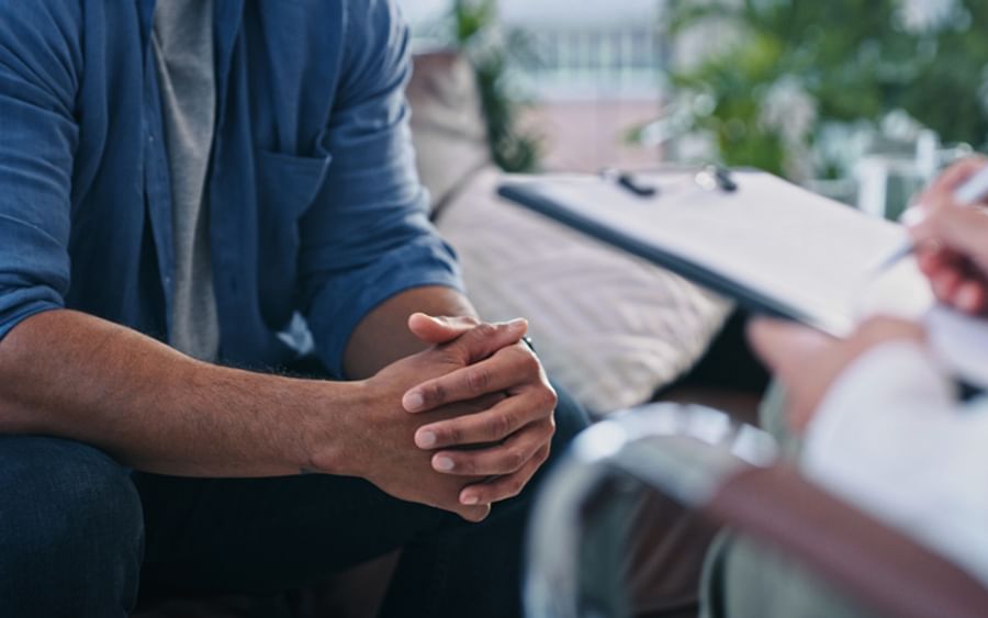 Person in blue shirt sits with clasped hands during a therapy session as another person takes notes.
