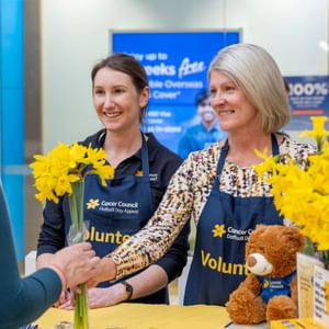 Two volunteers in blue aprons greet a customer, handing yellow daffodils at a Cancer Council stall.