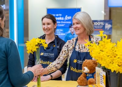 Two volunteers in blue aprons greet a customer, handing yellow daffodils at a Cancer Council stall.