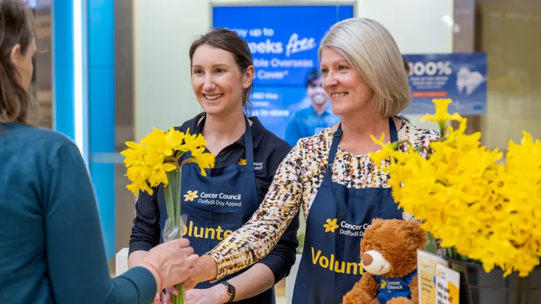 Two volunteers in blue aprons greet a customer, handing yellow daffodils at a Cancer Council stall.