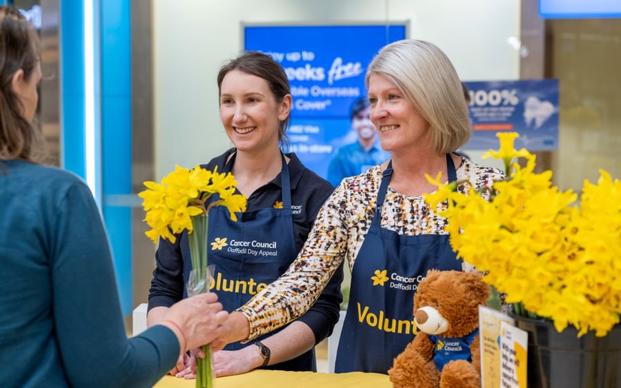 Two volunteers in blue aprons greet a customer, handing yellow daffodils at a Cancer Council stall.