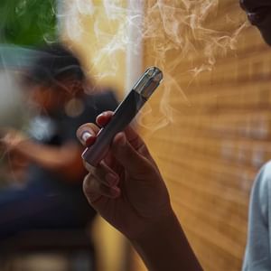 Close-up of a person holding a vape pen with smoke rising, brick wall in the background.