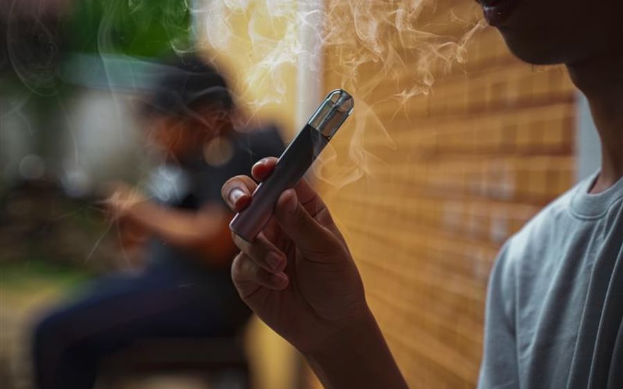 Close-up of a person holding a vape pen with smoke rising, brick wall in the background.