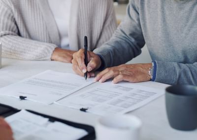 Two people sign documents at a white table with pens, papers and mugs.