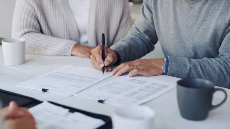 Two people sign documents at a white table with pens, papers and mugs.