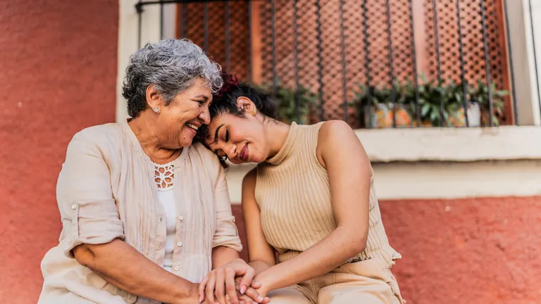 Two women outdoors, smiling and holding hands, one leaning on the other's shoulder.