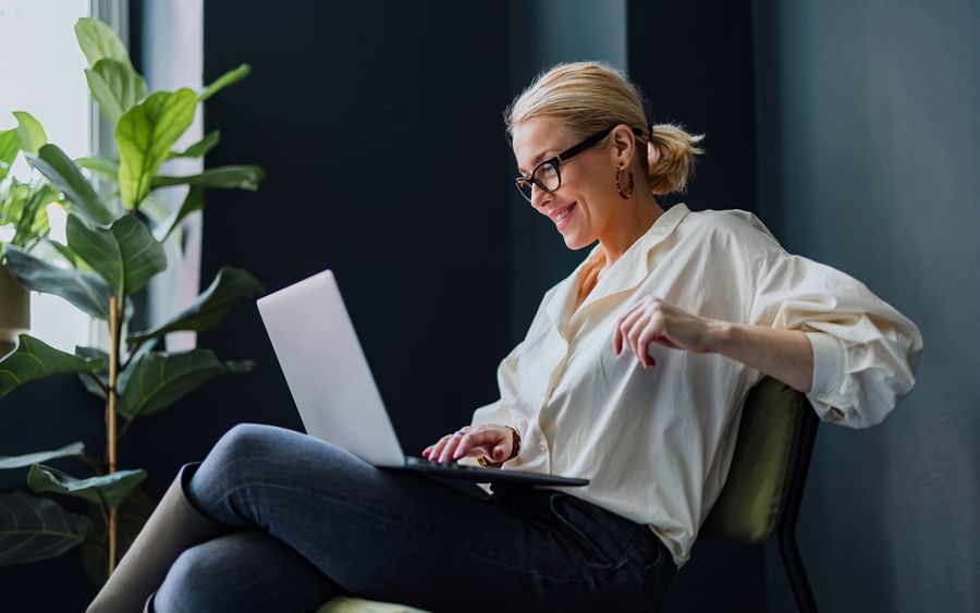 Woman sits side on with legs crossed on chair smiling at her laptop on her lap.