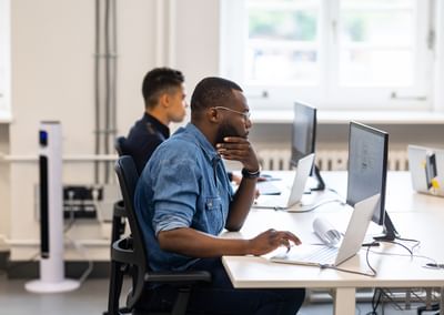 Person in a denim shirt using a laptop at a bright open-plan office; another person in background.