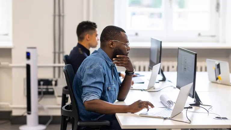 Person in a denim shirt using a laptop at a bright open-plan office; another person in background.