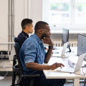 Person in a denim shirt using a laptop at a bright open-plan office; another person in background.