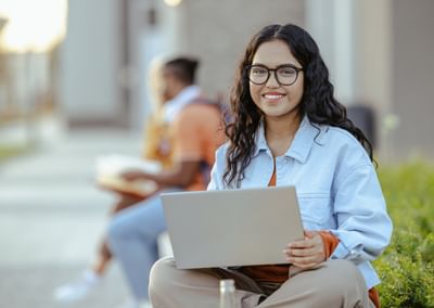 Person with glasses sits outdoors on campus, smiling, while holding a laptop.