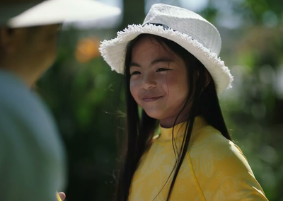 Young person with long hair, wearing a yellow patterned top and white fringed hat, smiling outdoors.