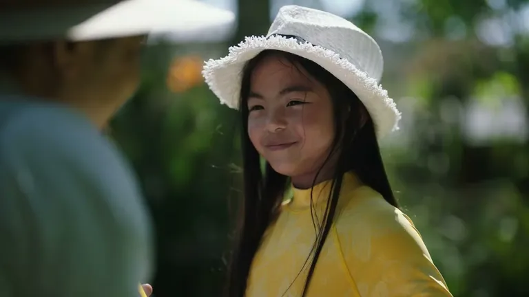 Young person with long hair, wearing a yellow patterned top and white fringed hat, smiling outdoors.
