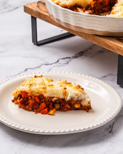 Lamb and Lentil Shepherd's Pie slice on a white plate. The remaining serving in a pie dish on a wooden board in the background.