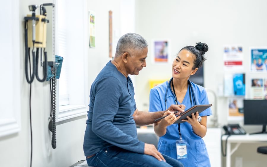 An elderly man sits in a hospital and talks with a female medical professional holding a clip board
