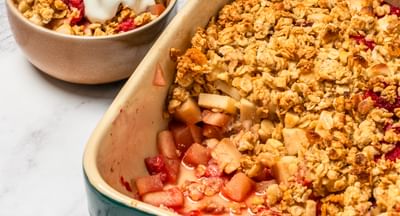 A green and white baking dish containing apple and raspberry crumble with a serving bowl on the side