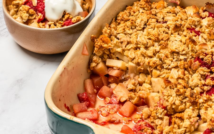 A green and white baking dish containing apple and raspberry crumble with a serving bowl on the side
