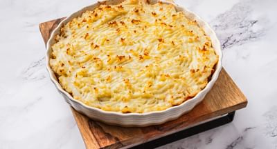 A baking dish filled with shepherd's pie on top of a square wooden chopping board