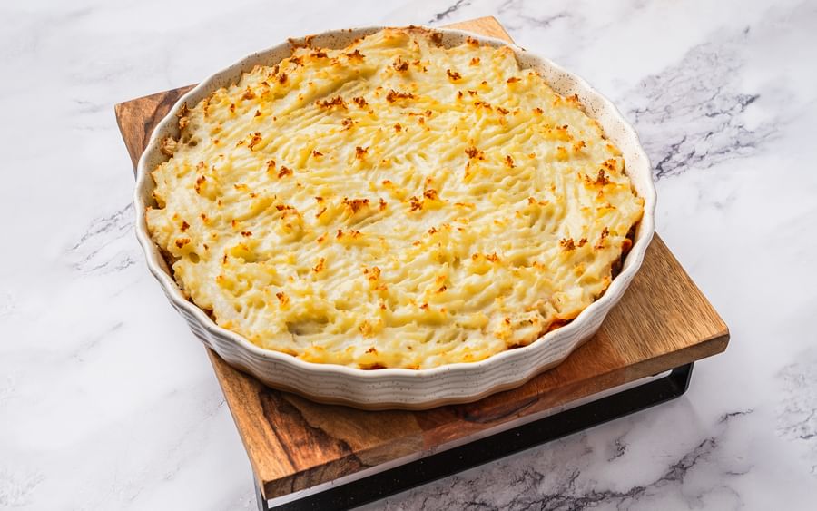 A baking dish filled with shepherd's pie on top of a square wooden chopping board