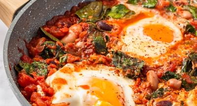 Close-up image of shakshuka, made using beans and eggs, in a grey frying pan on a marble surface
