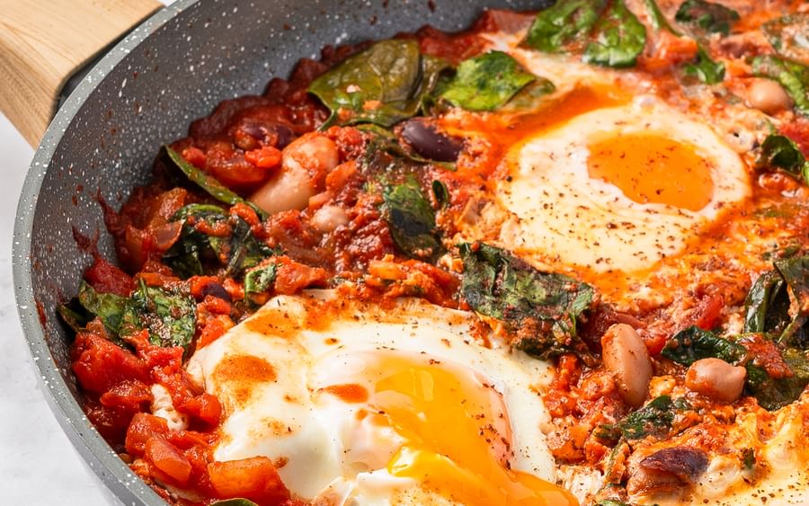 Close-up image of shakshuka, made using beans and eggs, in a grey frying pan on a marble surface