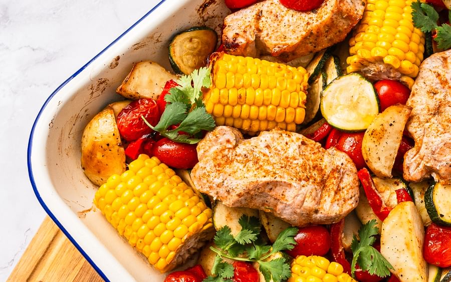 A white baking tray containing peri peri pork, corn, capsicum, cherry tomatoes and zucchini