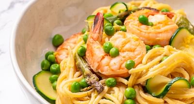 A bowl filled with spaghetti, prawns and green vegetables including peas, zucchini and asparagus