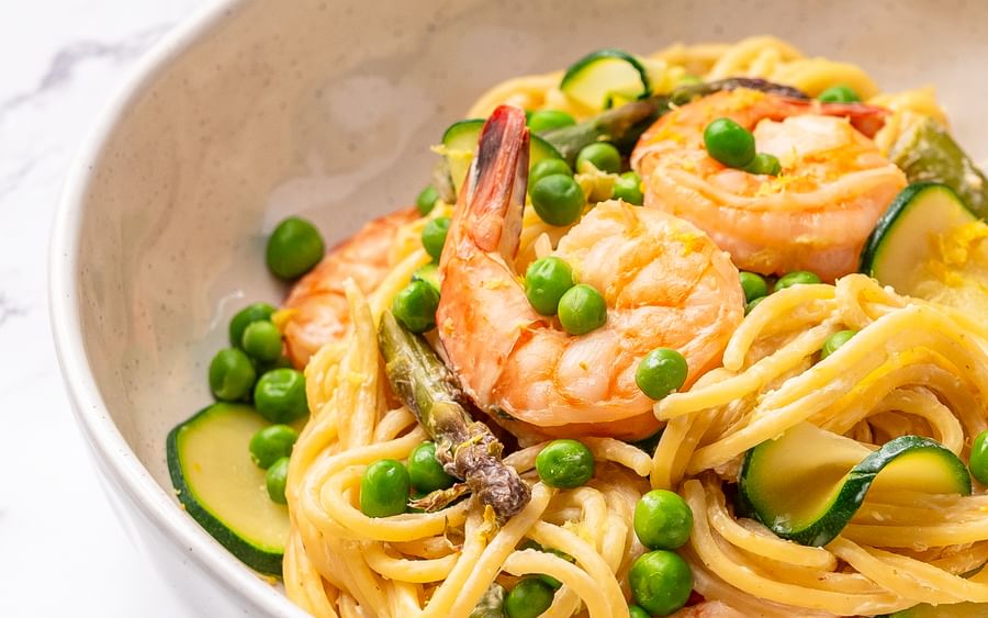 A bowl filled with spaghetti, prawns and green vegetables including peas, zucchini and asparagus