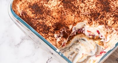 Creamy dessert with layers of berries and sponge and topped with cocoa powder; served in a square, glass dish sitting on top of a wooden chopping board with a handle; one serve of the dessert is in a small bowl.