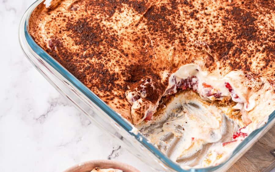 Creamy dessert with layers of berries and sponge and topped with cocoa powder; served in a square, glass dish sitting on top of a wooden chopping board with a handle; one serve of the dessert is in a small bowl.