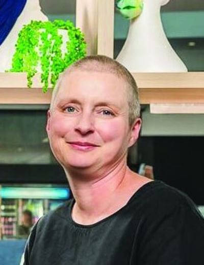 Person with shaved head sits on a light green sofa, wearing a black top, smiling; colourful wigs on display behind.