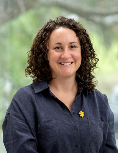 Smiling person with curly dark hair in a navy shirt, yellow flower pin, outdoors.