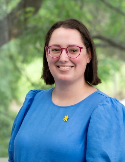Person with short dark hair and pink glasses, wearing a blue top with a yellow flower pin outdoors.