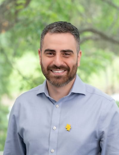 Person with short dark hair and beard, light blue shirt, yellow flower pin, smiling outdoors.