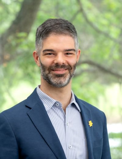 Smiling person in a navy blazer and striped shirt, yellow flower pin, outdoors with greenery.