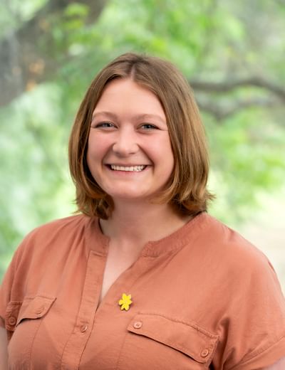 Smiling person with shoulder-length brown hair wearing an orange blouse with a yellow flower pin, outdoors.