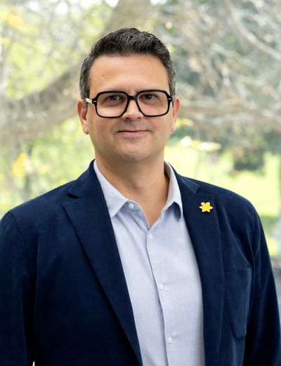 Person in a navy blazer and light shirt with glasses and a yellow lapel pin, outdoors.