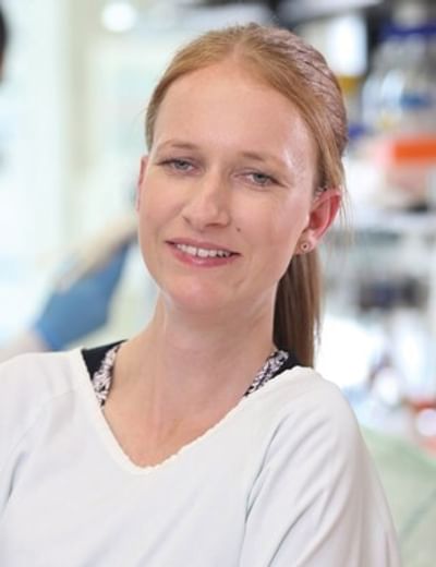 A smiling lab staff member with hair tied back in a white coat in a busy laboratory.