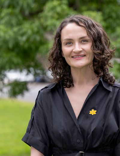 Person with curly brown hair in a black shirt with a yellow flower pin, smiling outdoors.