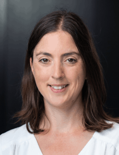 Portrait of a person with mid-length brown hair in a white blouse, smiling against a dark backdrop.