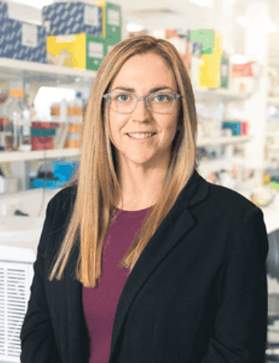 Person with shoulder-length hair and glasses wearing a blazer and maroon top, smiling in a bright store.