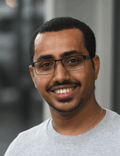 Smiling person with short hair, wearing glasses and a light grey shirt, outdoor urban background.