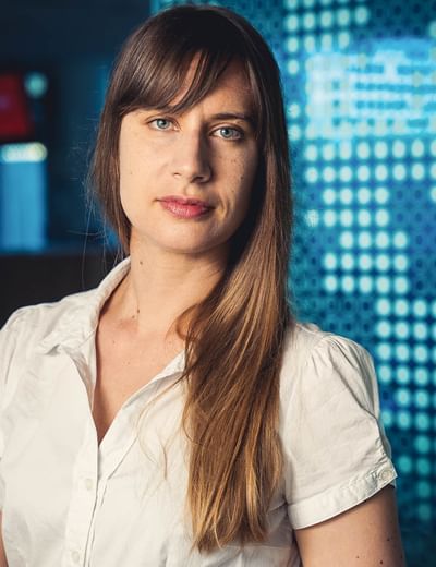 Person with long light brown hair in a white shirt, standing against a blue digital backdrop.